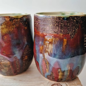 Könnte beinhalten: Zwei handgefertigte Keramikbecher mit einer einzigartigen Glasur. Die Becher weisen eine Mischung aus Farben auf, darunter tiefes Rot, Blau und Gold, wodurch ein strukturiertes, künstlerisches Aussehen entsteht. Die Ränder haben einen grünen Farbton.
