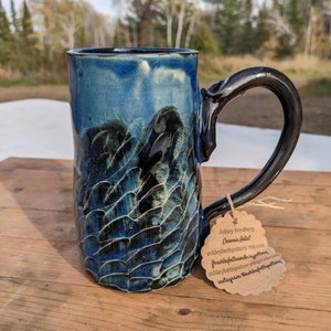 Puede incluir: Una taza de cerámica azul y negra con una superficie texturizada. La taza tiene una asa grande y está sentada sobre una mesa de madera. La taza tiene una etiqueta adjunta que dice "Ashley Nordberg Ceramic Artist ashleybethpottery.etsy.com facebook.com/ashleybethpottery ashleybethpottery@gmail.com instagram ashleybethpottery"