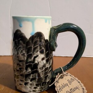 Puede incluir: Una taza de cerámica con un diseño texturizado en blanco y negro y un asa verde. La taza tiene un glaseado azul claro que gotea por el lado. La taza está sobre una superficie de madera con una etiqueta marrón que dice "Ashley Beth's Pottery".