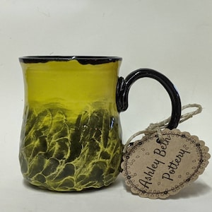 Puede incluir: Una taza de cerámica amarilla con un borde y un asa negros. La taza tiene un diseño texturizado de hojas en negro. Una etiqueta marrón con el texto "Ashley Beth Pottery" está unida al asa.