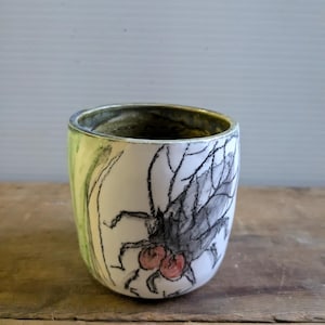 Puede incluir: Una taza de cerámica blanca con un esmalte verde y amarillo. La taza tiene un dibujo en blanco y negro de un insecto con puntos rojos.