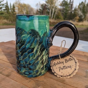 May include: A green and black ceramic mug with a textured surface and a black handle. The mug has a tag attached to it that says "Ashley Beth Pottery".