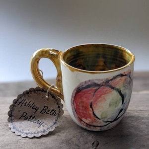 May include: A small, white ceramic mug with a brown handle and a colorful abstract design. The mug is sitting on a wooden surface. The mug has a tag that says "Ashley Beth Pottery".