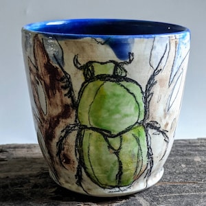 Puede incluir: Una taza de cerámica con un borde azul y un diseño pintado a mano de un escarabajo verde sobre un fondo blanco.