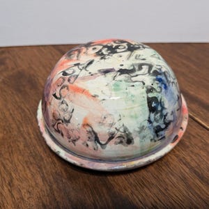 以下が含まれることがあります： 白いベースとカラフルな抽象的なデザインのセラミックドーム。デザインは、黒、ピンク、オレンジ、グリーン、ブルーのペイントの飛沫と渦巻きを特徴としています。
