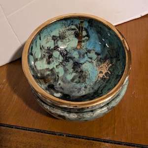 May include: A small, teal blue ceramic bowl with a gold rim and a black and gold design. The bowl has a textured surface and features a gold insect design.