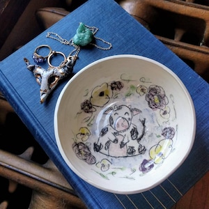 May include: A white ceramic bowl with a hand-painted design of a cow surrounded by flowers. The cow is black and white with a pink nose and is smiling. The flowers are yellow, purple, and green.