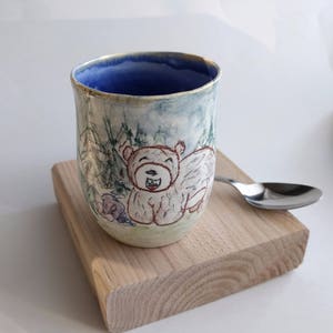 Op de afbeelding: Een keramische mok met een blauwe binnenkant en een handgeschilderd ontwerp van een beer in een boslandschap. De mok staat op een houten oppervlak.