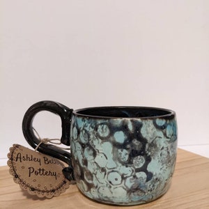 May include: A blue and black ceramic mug with a unique marbled glaze. The mug has a black handle and a tag that reads "Ashley Beth Pottery".