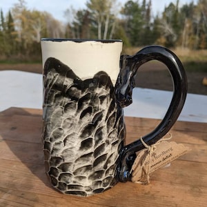 Puede incluir: Taza de cerámica negra y blanca con un diseño texturizado y abstracto. La taza tiene un asa negra y un interior blanco. La taza está sentada sobre una superficie de madera con una pequeña etiqueta que dice "Shelly Nordberg Ceramic Artist" y una dirección de sitio web.