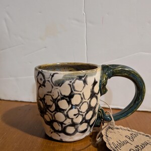 May include: A small, white ceramic mug with a black and white honeycomb pattern. The mug has a teal green handle and a brown tag that reads "Ashley Beth Pottery".