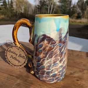 Puede incluir: Una taza de cerámica con un esmalte marrón, azul y blanco. La taza tiene un diseño único y texturizado y una asa grande. La taza está sentada sobre una superficie de madera. La taza tiene una etiqueta adjunta que dice "Ashley Beth Pottery".