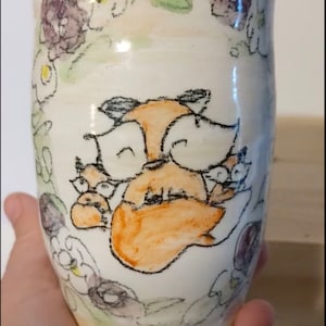 May include: A white ceramic mug with a hand-painted design of a fox with a floral border. The fox is orange and brown with black eyes and a black nose. The flowers are purple, yellow, and green.