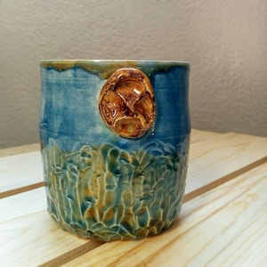 May include: A blue and brown ceramic mug with a textured surface and a raised, brown, oval-shaped design on the side.