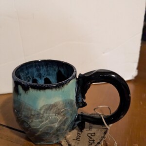 Puede incluir: Una taza de cerámica azul y negra con una forma única e irregular y un asa negra. La taza tiene un esmalte moteado y una base azul claro. La taza está sobre una superficie de madera con una etiqueta que dice "Casey Beth Pottery".