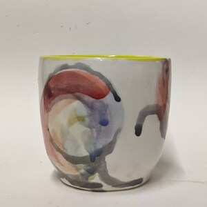 May include: A white ceramic mug with a yellow rim and abstract watercolor-like designs in shades of red, blue, and green.