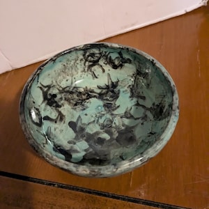 May include: A teal ceramic bowl with a black and white marbled design. The bowl has a rounded shape and a smooth, glossy finish.