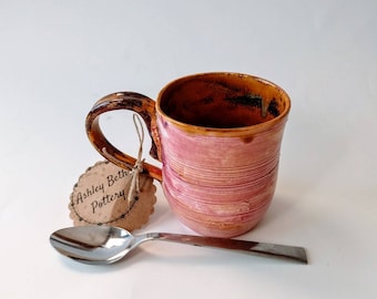 May include: A handmade ceramic mug with a pink and orange glaze. The mug has a brown handle and a small tag that says "Ashley Beth Pottery".