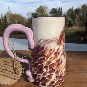 Puede incluir: Una taza de cerámica con asa morada y cuerpo con estampado marrón y blanco. La taza está sobre una superficie de madera. La etiqueta dice "Ashley Nobe Ceramic Art" e incluye información de contacto.