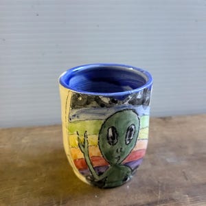 May include: A small ceramic mug with a hand-painted design of an alien waving with a rainbow background. The mug has a blue rim and a white base.