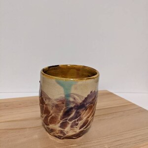 Puede incluir: Una taza de cerámica con un esmalte texturizado marrón y amarillo. La taza tiene un interior marrón claro y un detalle de esmalte azul claro.