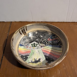 May include: A ceramic bowl with a hand-painted design of a UFO with a rainbow and stars. The bowl has a gold rim and is perfect for holding jewelry or other small items.