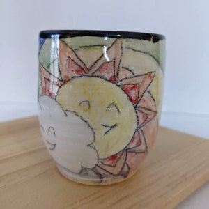 Könnte beinhalten: Eine weiße Keramiktasse mit handgemaltem Design einer Sonne und einer Wolke. Die Sonne ist gelb mit einem lächelnden Gesicht und die Wolke ist weiß mit einem lächelnden Gesicht. Der Hintergrund ist hellgrün mit roten und orangen Akzenten.