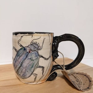 Peut inclure: Une tasse en céramique avec une anse noire et un motif peint à la main d'un scarabée bleu sur fond blanc. La tasse est recouverte d'un glaçage brun et repose sur une surface en bois. La tasse a une étiquette attachée à la poignée qui dit "Artisanat".