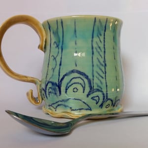 Może przedstawiać: Jasnoniebieski kubek ceramiczny z żółtym uchwytem i wzorem w kolorze niebieskim i zielonym. Kubek stoi na srebrnej łyżce.