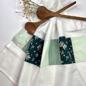 May include: White kitchen towel with a decorative band featuring floral patterns in shades of green and white. Two wooden spoons rest on the towel. The towel has a textured, ribbed appearance.