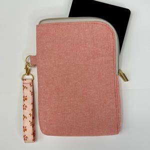 May include: A blush pink zippered case with a gold-toned zipper and hardware. A black object is partially visible inside the case. A floral patterned wrist strap is attached to the case with a gold clasp. The case is made of a textured fabric.