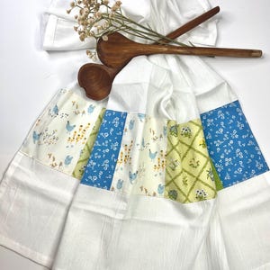 May include: White kitchen towels with patterned panels featuring chickens, flowers, and blue floral designs. Two wooden spoons and dried flowers rest on top. The towels are folded and arranged on a white surface.