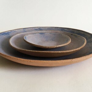 May include: Three ceramic plates nested together. The largest plate is a light brown color with a blue glaze. The middle plate is a darker brown color with a blue glaze. The smallest plate is a light purple color with a blue glaze.