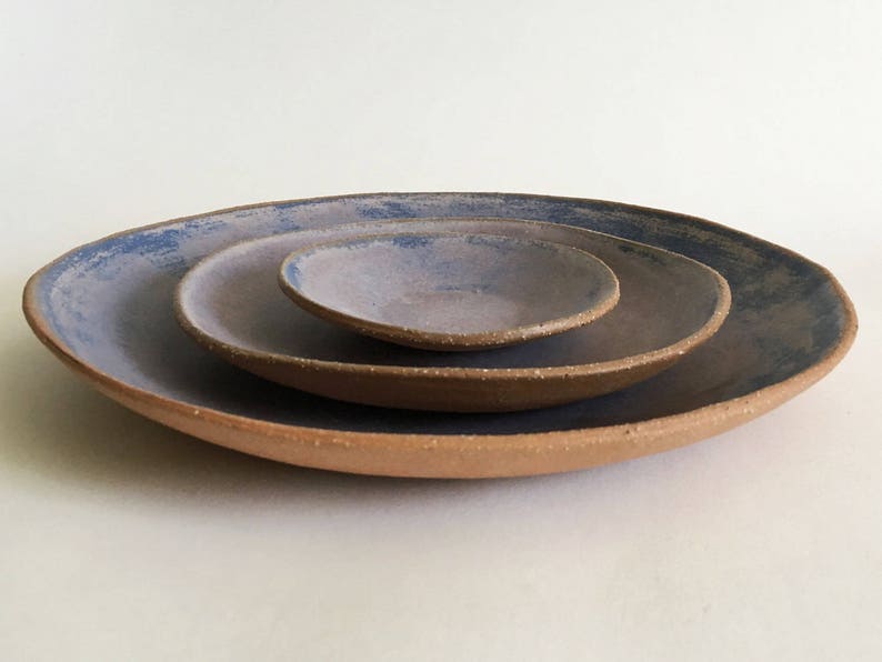 May include: Three ceramic plates nested together. The largest plate is a light brown color with a blue glaze. The middle plate is a darker brown color with a blue glaze. The smallest plate is a light purple color with a blue glaze.