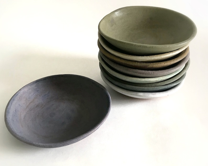 Puede incluir: Un conjunto de peque&ntilde;os cuencos de cer&aacute;mica hechos a mano en varios colores apagados, incluyendo verde, marr&oacute;n y gris. Un cuenco est&aacute; aislado en primer plano, en un color gris azulado oscuro. Los cuencos tienen una forma org&aacute;nica, ligeramente irregular.