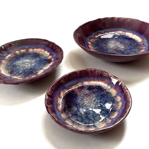 May include: Set of three ceramic bowls with a purple, blue, and white glaze. The bowls have a wavy edge and a unique, iridescent finish.