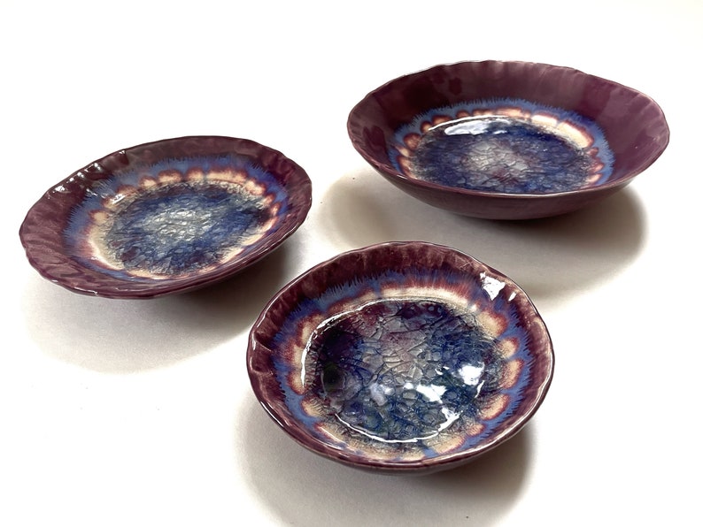 May include: Set of three ceramic bowls with a purple, blue, and white glaze. The bowls have a wavy edge and a unique, iridescent finish.
