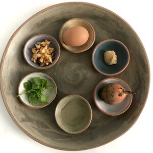 May include: A brown ceramic plate with six smaller bowls arranged in a circle. The bowls contain a hard-boiled egg, chopped apples, parsley, horseradish, a beet, and a small bowl of water.