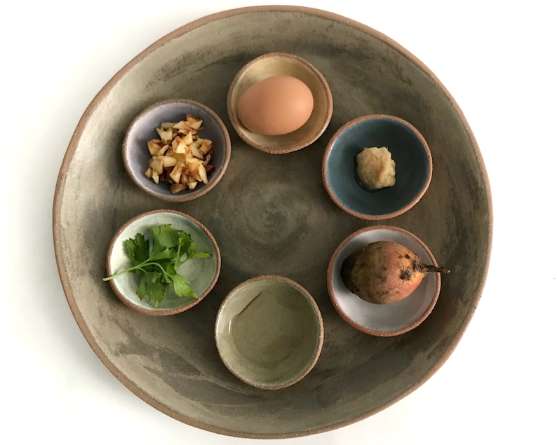May include: A brown ceramic plate with six smaller bowls arranged in a circle. The bowls contain a hard-boiled egg, chopped apples, parsley, horseradish, a beet, and a small bowl of water.