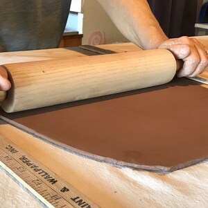 May include: A person rolling out a slab of brown clay with a wooden rolling pin on a white surface. A ruler is visible in the foreground.