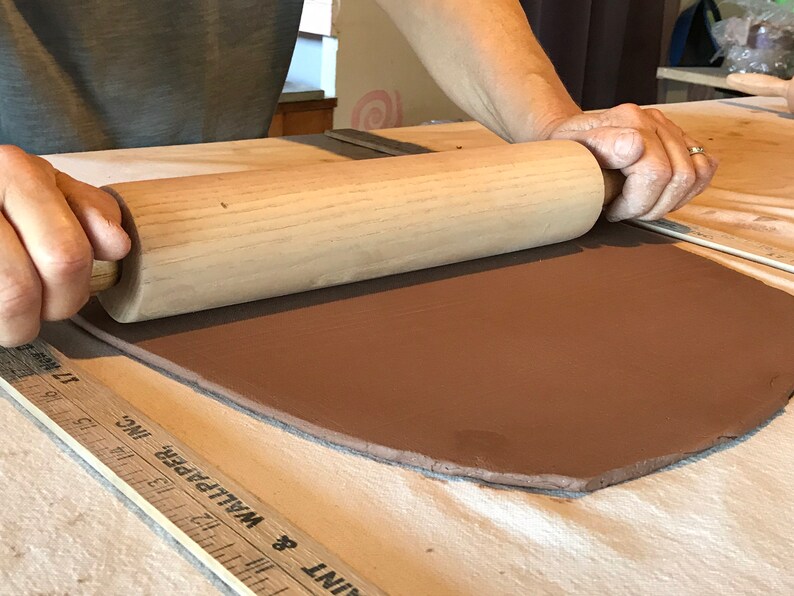 May include: A person rolling out a slab of brown clay with a wooden rolling pin on a white surface. A ruler is visible in the foreground.