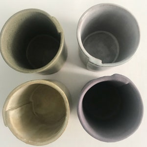 May include: Four ceramic cups in different shades of green, grey, and purple. The cups are all cylindrical and have a slightly flared lip.