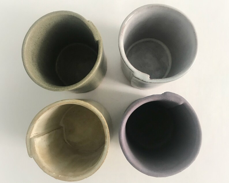 May include: Four ceramic cups in different shades of green, grey, and purple. The cups are all cylindrical and have a slightly flared lip.