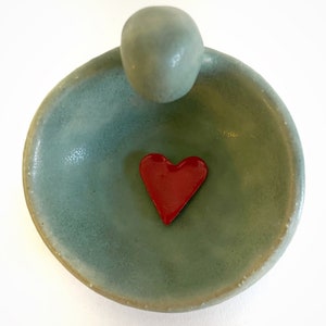 May include: A small, light blue ceramic dish with a rounded, raised edge. A red heart-shaped object is centered in the dish.