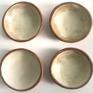 May include: Four small, round, terracotta-colored bowls with a white glaze inside. The bowls are arranged in a square pattern on a white surface.