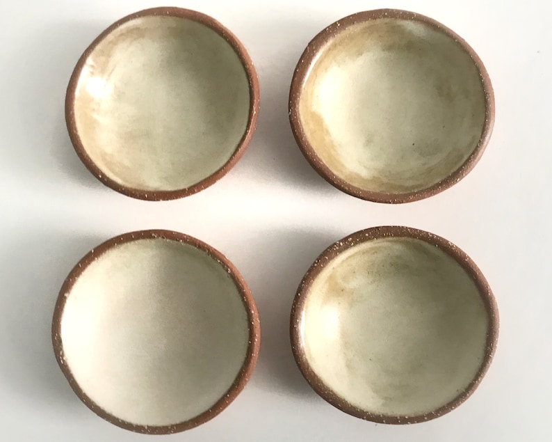May include: Four small, round, terracotta-colored bowls with a white glaze inside. The bowls are arranged in a square pattern on a white surface.