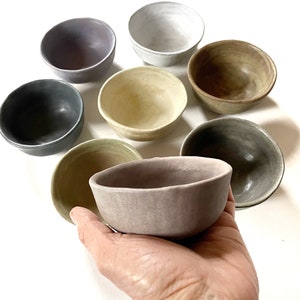 May include: A collection of seven small ceramic bowls in various colors, including gray, brown, beige, and purple. The bowls are arranged in a circle on a white surface, with one bowl being held in a hand.