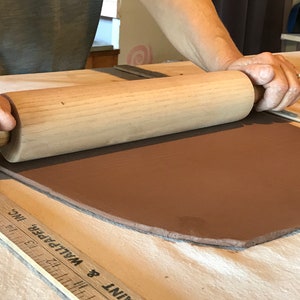 May include: A person rolling out a slab of brown clay with a wooden rolling pin on a wooden surface. A ruler with the text "PAINT & WALLPAPER INC." is visible in the foreground.