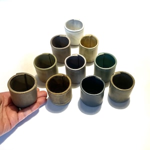 Può includere: Dieci piccole tazze in ceramica in varie tonalità di verde, marrone e grigio. Le tazze sono disposte a forma di triangolo su una superficie bianca.