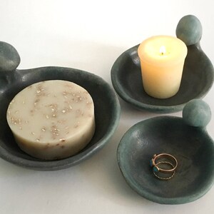 May include: Three ceramic dishes with a unique, organic shape. The dishes are in shades of teal and grey. One dish holds a bar of soap, another holds a candle, and the third holds a ring.